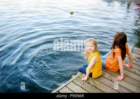 Deux mignonnes petites filles assis sur une plate-forme de bois par la rivière ou lac trempant leurs pieds dans l'eau chaude journée d'été sur Banque D'Images