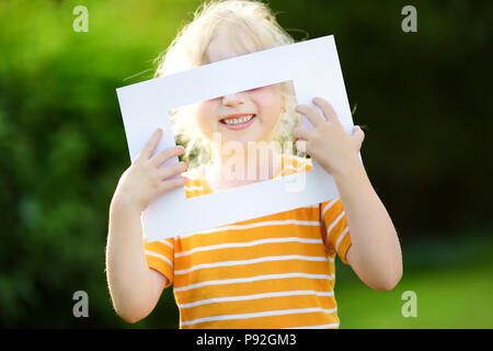 Joyeux cute little girl holding cadre photo blanc en face de son visage. L'encadrement de l'enfant adorable son visage souriant. Banque D'Images