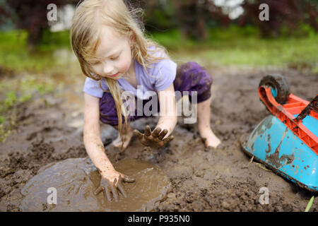 Funny little girl playing dans une grande flaque de boue humide par beau jour d'été. Enfant de se salir en creusant dans le sol boueux. Jeux en plein air. malpropre Banque D'Images