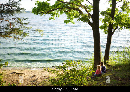 Deux mignonnes petites soeurs assis au bord du lac avec sa vue sur le coucher de soleil. Les enfants d'explorer la nature. Activités en famille l'été. Banque D'Images