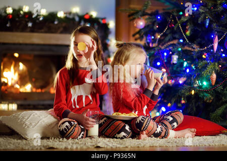 Happy Little Sisters ayant du lait et les cookies par une cheminée dans un confortable salon sombre la veille de Noël. La célébration de Noël à la maison. Banque D'Images