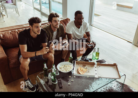 Les amis s'amusant avec des boissons et des collations tout en jouant le jeu vidéo à la maison. Deux hommes jouer jeu vidéo joysticks holding tandis qu'un autre homme est titulaire d'un mob Banque D'Images