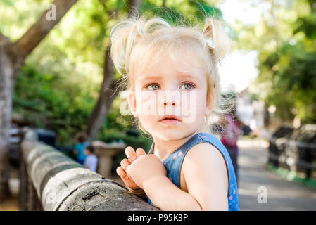 Close up portrait of cute peu désorientés blondy bébé fille à côté et s'appuyant sur une clôture en bois dans le zoo ou parc de la ville enfant , sécurité Banque D'Images