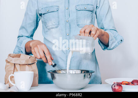 Jeune femme la cuisson gâteau aux prunes, en fouettant la pâte à table debout sur fond blanc. Les mains de femmes de verser le lait dans un bol en métal à côté d'un sac en papier o Banque D'Images