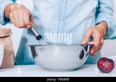 Close-up young woman's hands holding fouetter et bol en métal tout en faisant un gâteau aux fruits. Pâte à tarte cuisson femelle sur le tableau blanc du papier près de sac de farine Banque D'Images