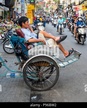 Un homme assis sur un pousse-pousse près d'une route à fort trafic de motos, motos et scooters à Ho Chi Minh City, Vietnam. Banque D'Images