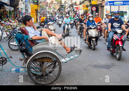 Un homme assis sur un pousse-pousse près d'une route à fort trafic de motos, motos et scooters à Ho Chi Minh City, Vietnam. Banque D'Images