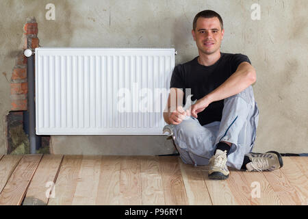 Jeune beau plombier assis sur marbre avec une clé dans la main près du radiateur de chauffage installé avec succès en salle vide d'un appartement nouvellement construit ou h Banque D'Images