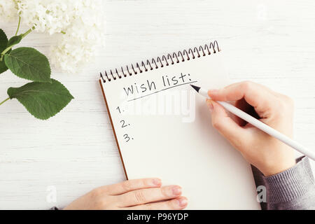 Woman's hand writing liste de souhaits dans l'ordinateur portable sur une table en bois blanc. Lieu de travail Planification et concept, vue d'en haut. Banque D'Images