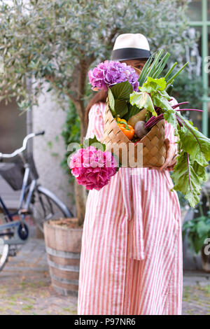 Une fille dans un chapeau sur une rue de la ville avec un grand panier de légumes et de fleurs dans ses mains. Belle femme avec des boutiques et un vélo. Marcher le long des rues. Copy space Banque D'Images