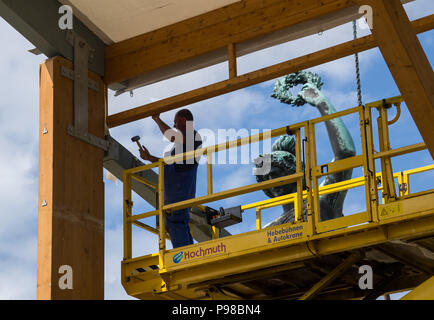 Munich, Allemagne. 16 juillet, 2018. Un travailleur fixation de la poutre du toit à poutres en bois la structure d'un chapiteau. Le Bavaria statue peut être vu dans l'arrière-plan. Cette année, l'Oktoberfest se déroule du 22 septembre au 07 octobre 2018. Crédit : Peter Kneffel/dpa/Alamy Live News Banque D'Images