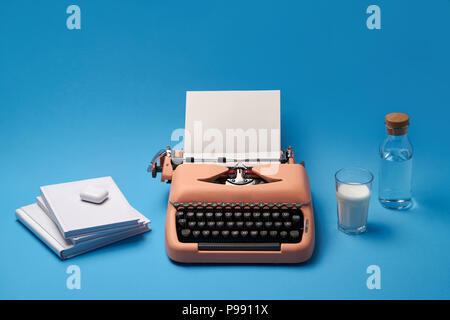 Machine à écrire vintage avec une feuille de papier sur le fond bleu du studio. À côté il y a peu de livres, verre de lait et peu de bouteille avec une c Banque D'Images