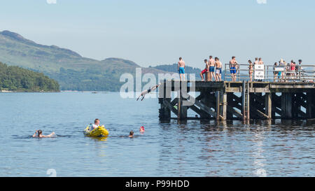 Les jeunes bénéficiant d'une chaude journée à Luss, Loch Lomond - natation, canotage et de plongée sous-marine au large de la jetée au cours de la canicule de Juin/Juillet 2018 Banque D'Images