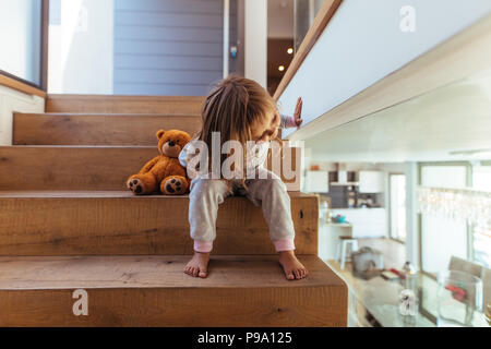 Cute little girl sitting on escalier et à la recherche à travers un mur de verre à l'intérieur. Petite fille avec des pics de salle de séjour à la maison. Escalier en bois Banque D'Images