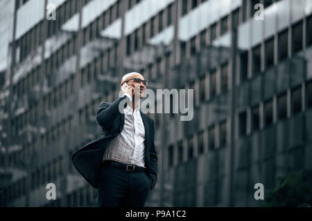 Smiling businessman de conversations au téléphone cellulaire extérieur permanent avec une main dans la poche. L'homme à marcher sur les vêtements officiels street talking on mobile phone. Banque D'Images