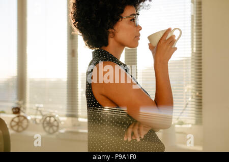 Vue latérale du young businesswoman in casuals drinking coffee in office. Femme penser à quelque chose alors que le café pendant le travail. Banque D'Images