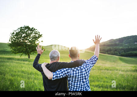 Un adulte avec des fils hipster père sur une promenade dans la nature au coucher du soleil, les bras autour de l'autre. Banque D'Images