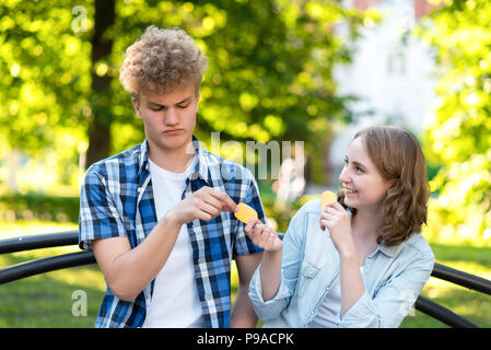 De jeunes étudiants en été dans un parc à l'extérieur. Ils sont assis sur un banc dans la ville. Fille et happy smiling donne de la nourriture à guy. Le mec est gêné. Le concept de la méfiance. L'émotion de bonté. Banque D'Images