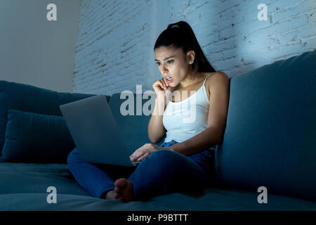 Belle jeune femme hispanique addict internet en jeans et débardeur sur le canapé surprise face à l'expression avec ordinateur portable tard dans la nuit à l'aide de chat Banque D'Images