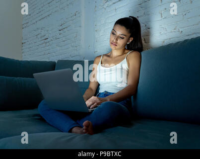 Belle jeune femme addict internet hispanique dans les tenues de assis sur le canapé et s'ennuie de travail avec ordinateur portable assez tard dans la nuit à da Banque D'Images