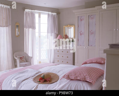 Chapeau de paille et rose vérifié coussins sur lit dans une chambre avec gris pâle voile blanc rideaux sur les fenêtres Banque D'Images