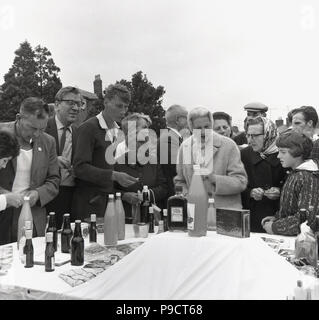 1967, anglais Fête du village, l'assortiment des populations locales se réunissent à l'extérieur autour d'une table à la tombola au prix à gagner, y compris des bouteilles de jus de pomme, bière, spiritueux et une boîte de chocolats à la magie noire, England, UK. Banque D'Images