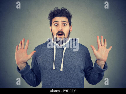 Jeune homme barbu in hoodie à avec la peur et l'étonnement à l'appareil photo avec les mains sur fond gris Banque D'Images