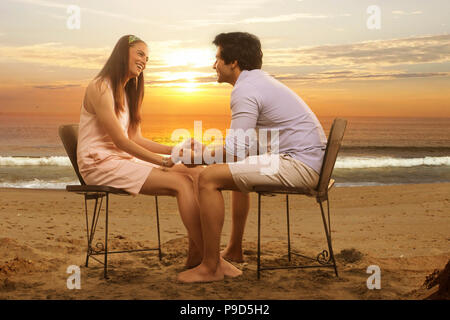 Jeune couple assis dans des chaises holding hands on beach at sunset Banque D'Images