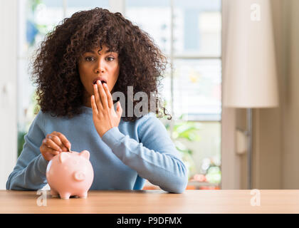 African American Woman fait économiser de l'argent dans la tirelire couvrir la bouche à part choqué avec honte pour erreur, expression de la peur, peur en silence, secret c Banque D'Images