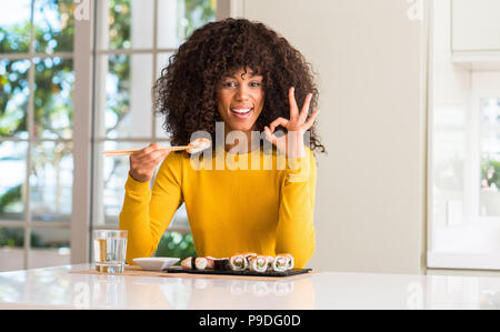African American Woman eating sushi à l'aide de baguettes à la maison faisant signe ok avec les doigts, symbole excellent Banque D'Images