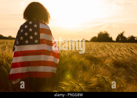 Mixed Race African American girl adolescent femme jeune femme dans un champ de blé ou d'orge enveloppés dans USA stars and stripes flag dans golden sunse Banque D'Images