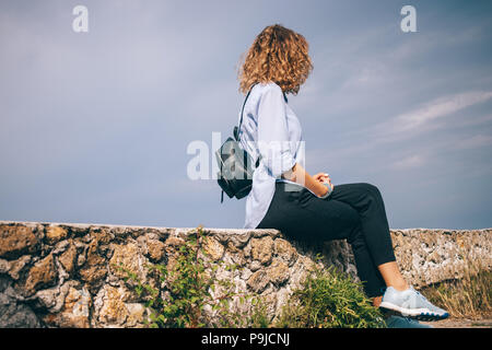 Femme portant une chemise bleue, un pantalon noir et sac à dos, assis sur de vieux parapet. Jeune femme avec des cheveux bouclés en appui à l'extérieur et regarde ailleurs. Banque D'Images