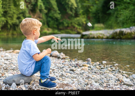 Enfant seul avec des cheveux blonds, jette une pierre sur la rive de la rivière,journée ensoleillée,photo horizontale Banque D'Images