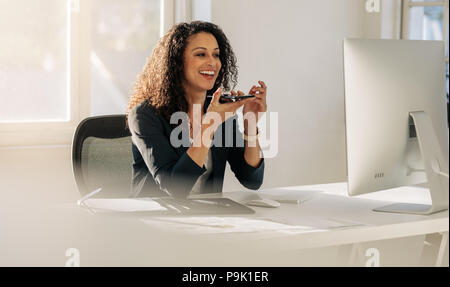 Smiling woman entrepreneur à son bureau à l'office de tourisme de conversations au téléphone mobile sur l'Orateur. Femme assise en face de computer in office talking on cell ph Banque D'Images