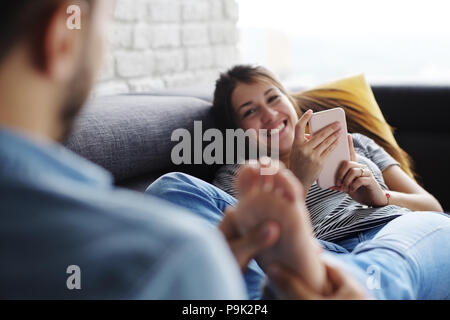 Young hispanic couple portant sur la table à la maison, à l'aide d'un téléphone mobile pour l'Internet et les médias sociaux. Le petit ami est de donner un massage de pieds de la wo Banque D'Images