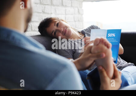 Young hispanic couple portant sur la table à la maison, tenant un billet circulaire et de la planification de mon prochain voyage. Le petit ami est de donner un massage de pieds de la femme. Banque D'Images
