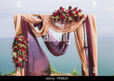 Cérémonie de mariage style Boho avec vue sur la mer. Décorées avec des roses rouges, jaunes, orchidées et de raisin pourpre et le tissu beige Banque D'Images