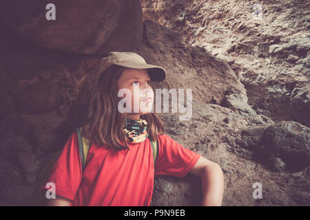 Une petite fille regarde pensivement au milieu d'une longue randonnée, à l'abri sous les rochers à flanc de montagne. Elle porte un foulard uv, casquette, crème solaire. Banque D'Images