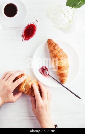 Café, croissants, confiture sur la table en bois blanc. Petit-déjeuner traditionnel français. Woman's hands holding croissant. Vue d'en haut. Banque D'Images