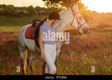 Prendre soin des animaux, l'amour et l'amitié concept. Jeune Jockey girl petting et serrant cheval blanc dans le coucher du soleil du soir. Serveur Sun flare Banque D'Images