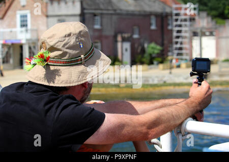 Bancs, Dorset, Angleterre - 02 juin 2018 : Un homme dans un floppy hat tournage vidéo sur un appareil photo d'action qu'un bateau arrive dans un petit port Banque D'Images