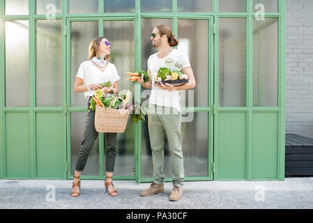 Jeune couple végétariens avec sac et boîte pleine de produits crus frais provenant du marché local en plein air sur le fond vert Banque D'Images