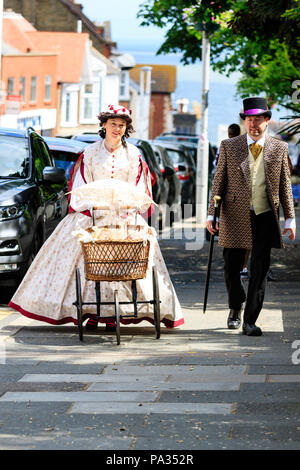 Jeune couple habillés en costume victorien, marcher le long de la rue ensoleillée la pram, poussant vers l'afficheur. Une partie de la semaine Charles Dickens festival, Broadstairs Banque D'Images