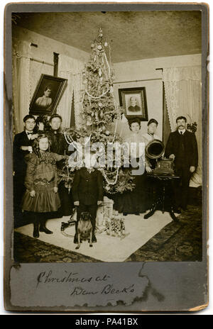 . Anglais : Vertical, sépia photographie d'une famille réunis autour d'un arbre de Noël dans leur maison. Un adolescent, une jeune fille et une femme se tenir ensemble à gauche sous un portrait d'une femme. Un jeune garçon et le chien se tenir en face d'un l'arbre et quelques cadeaux dont un train set et soldats de plomb. À droite sont une deux femmes et un homme réunis autour d'un phonographe. L'arbre est haut et étroit et est décorée avec des ornements en verre-ball, des bougies, des chiffres, des perles, et une chaîne de popcorn. Titre : 'Noël à Dan Beck's.' . vers 1905 2 "Noël à Dan Beck's." Banque D'Images