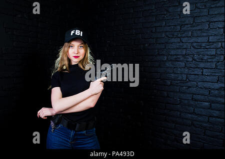 FBI agent féminin en cap et à canon au studio contre un mur de briques sombres. Banque D'Images