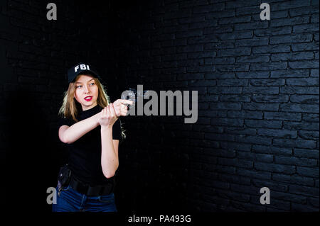 FBI agent féminin en cap et à canon au studio contre un mur de briques sombres. Banque D'Images