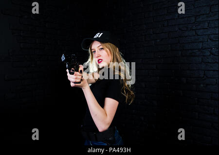 FBI agent féminin en cap et à canon au studio contre un mur de briques sombres. Banque D'Images