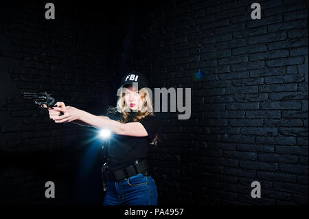 FBI agent féminin en cap et à canon au studio contre un mur de briques sombres. Banque D'Images