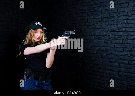 FBI agent féminin en cap et à canon au studio contre un mur de briques sombres. Banque D'Images