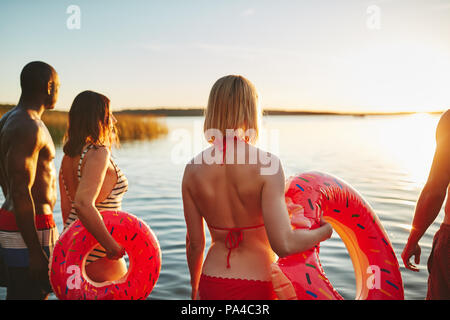 Coquille d'un groupe de jeunes amis diversifié en maillot de bain dans un lac permanent en regardant le coucher du soleil ensemble Banque D'Images